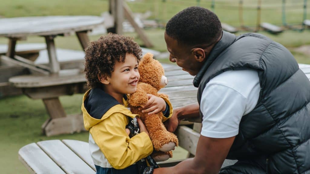 A Black grownup speaks to a small child embracing a teddy bear.