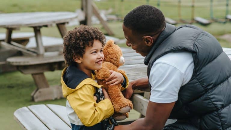 A Black grownup speaks to a small child embracing a teddy bear.