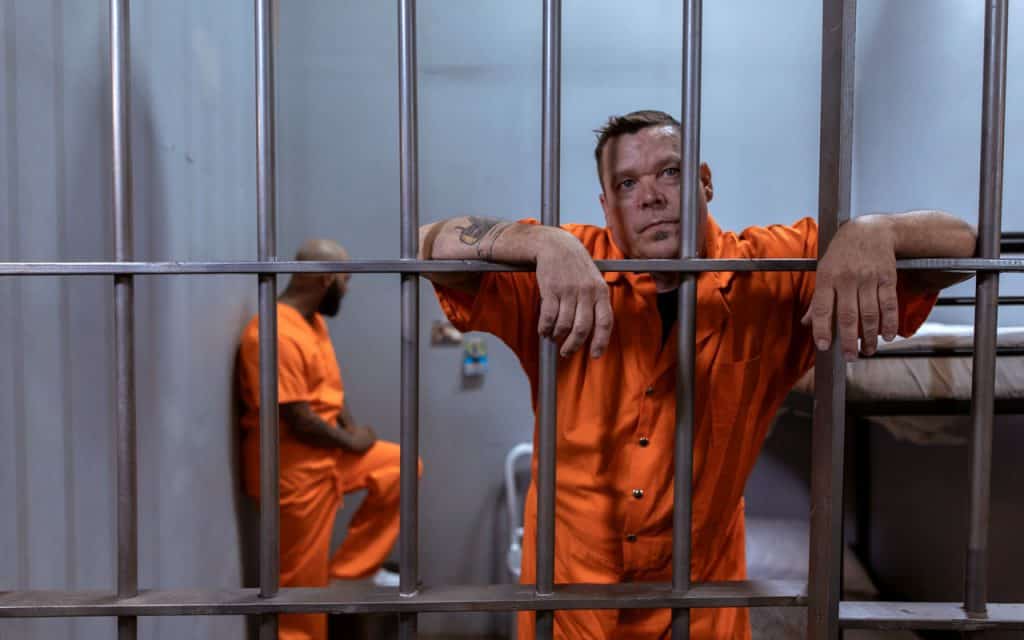 Two masc-presenting people in orange jumpsuits sit in a prison cell.
