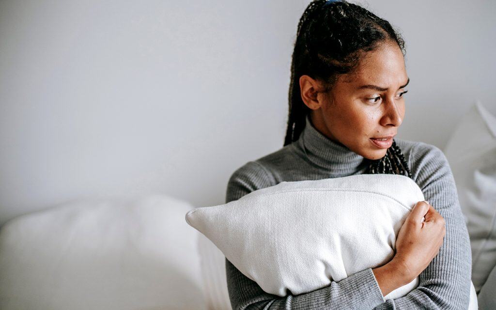 Black women a concerned expression sitting on a bed clutching a pillow to her chest.