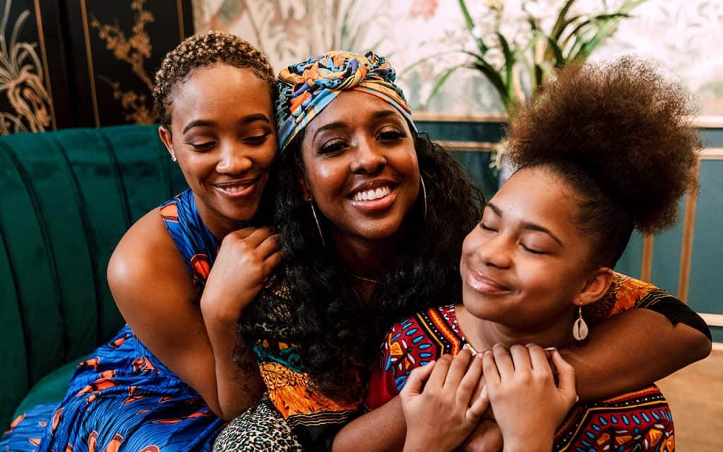 Three black women embracing and smiling in bright patterned clothing.