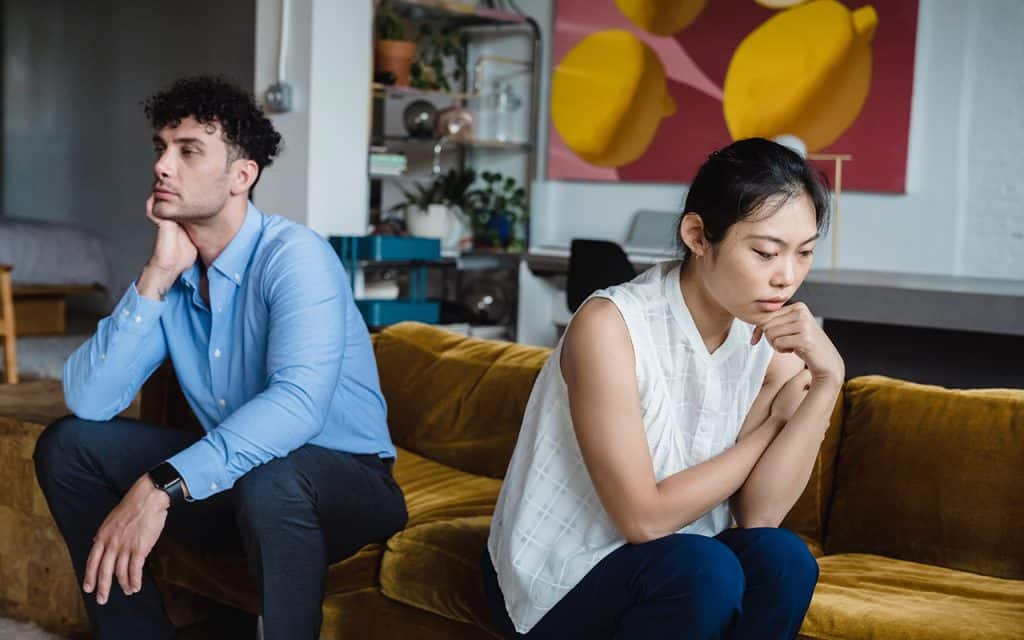 Couple with upset expressions sit on a couch facing away from each other