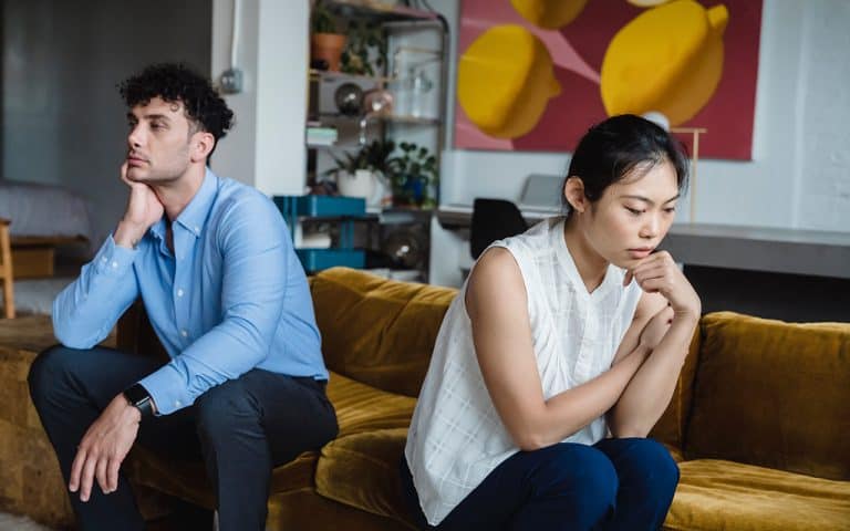 Couple with upset expressions sit on a couch facing away from each other