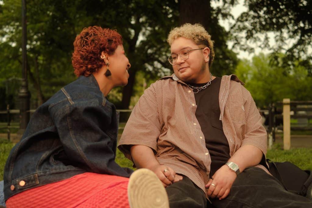 Two gender-non-conforming people sit together outside. They are looking towards each other and smiling.