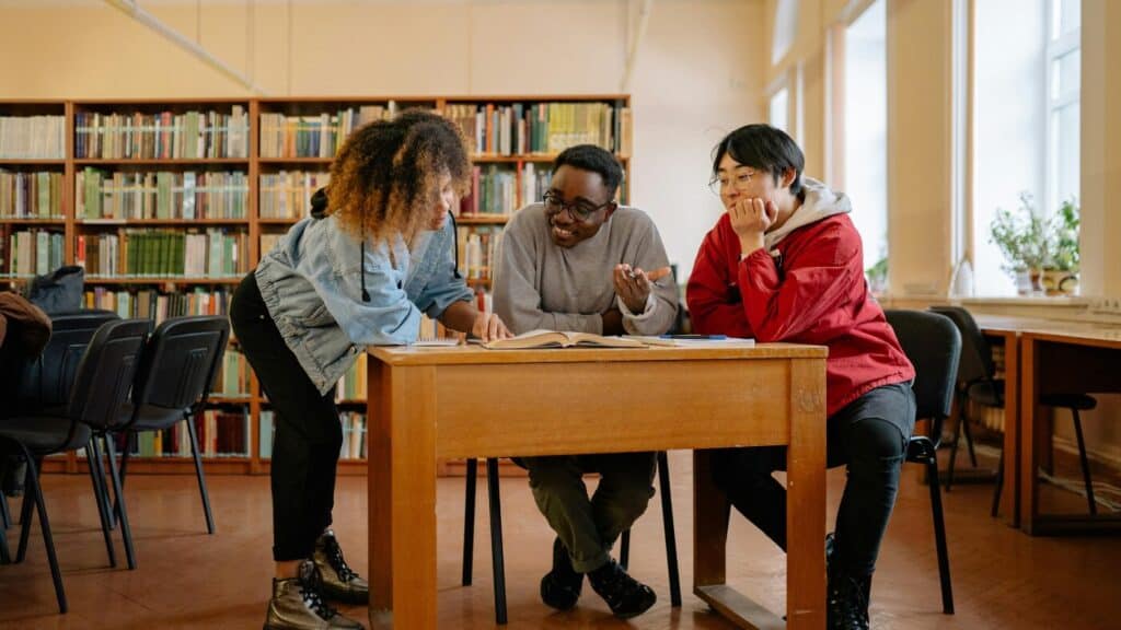 Three students work together at a desk in a library, smiling and talking.