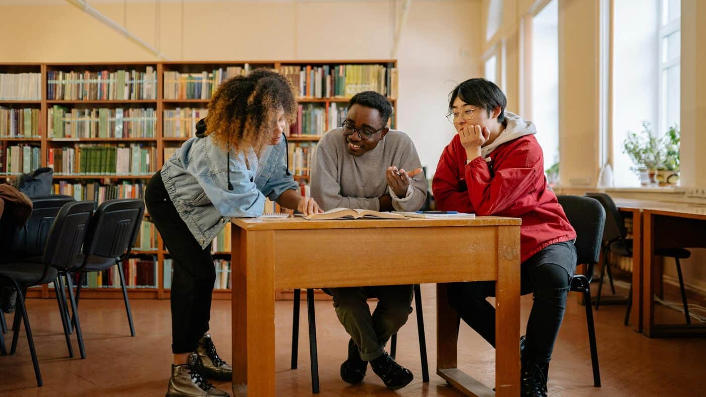 Three students work together at a desk in a library, smiling and talking.