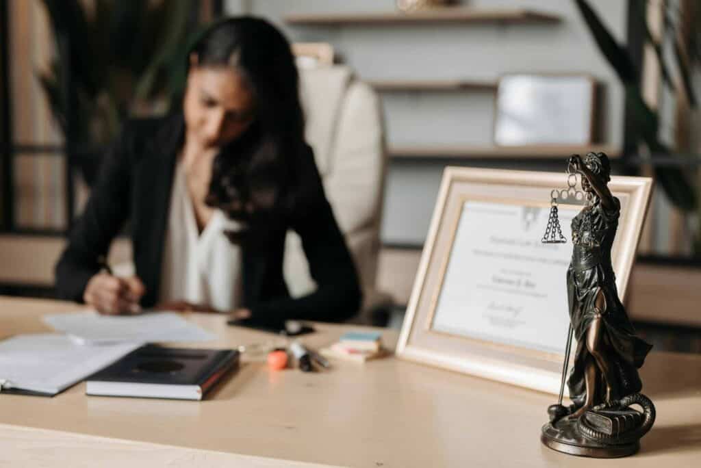 A woman works at a desk. In the foreground of the photo is a small figure of a woman holding the "scales of justice".