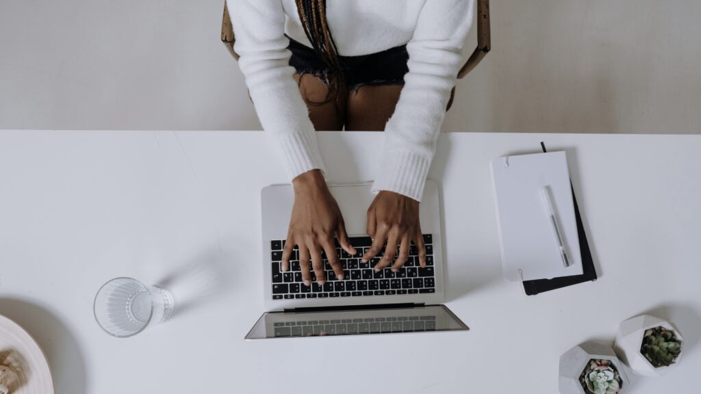 An overhead photo of a Black person typing on a laptop, with notes and a glass of water near them.