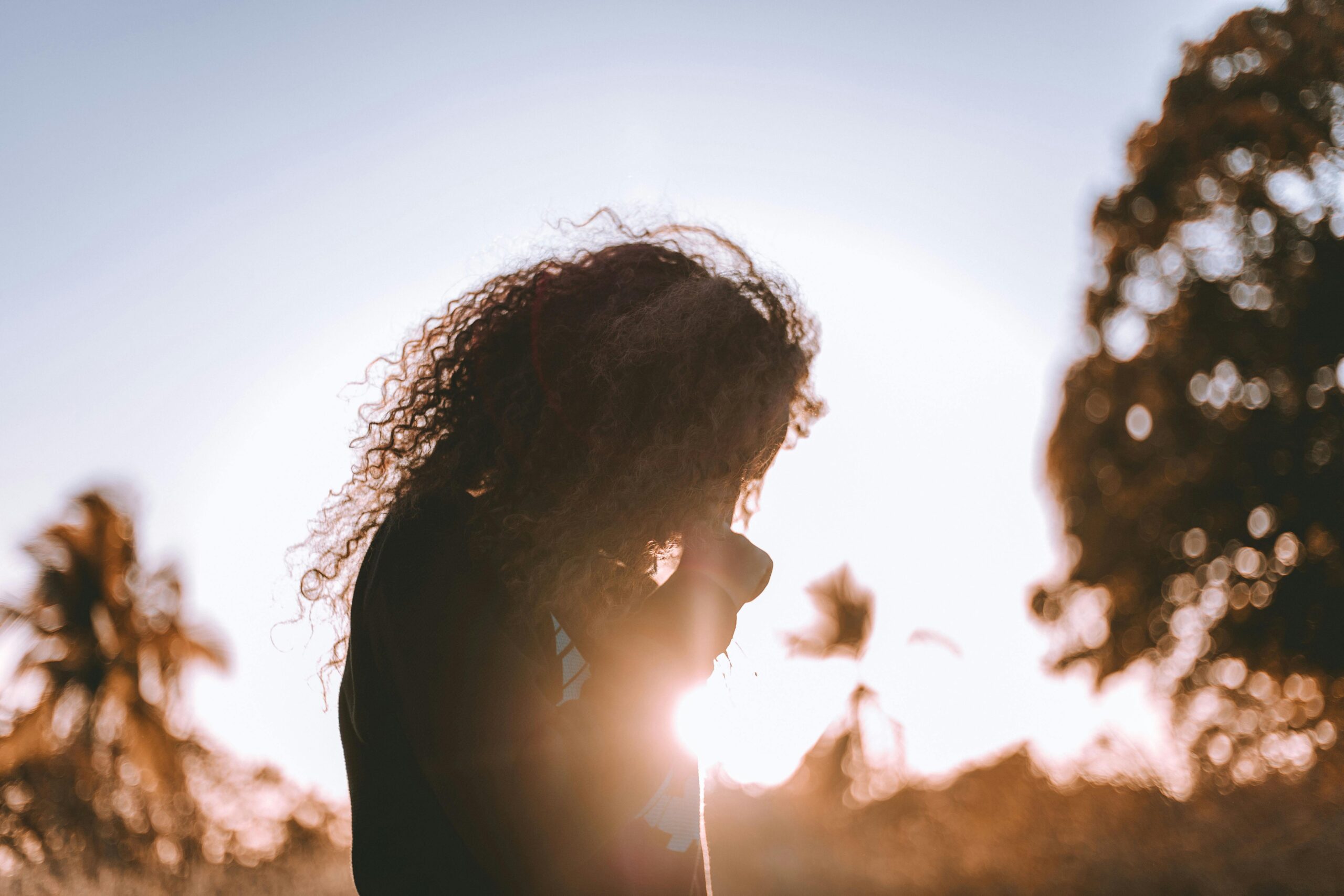 Woman with curly hair stands outside in front of the sun, looking down.