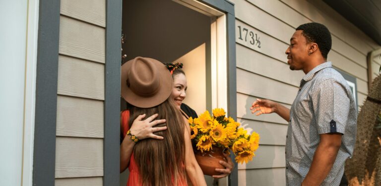 One person stands inside a doorway, hugging one person in a hat, and looking towards another person standing in front of them.