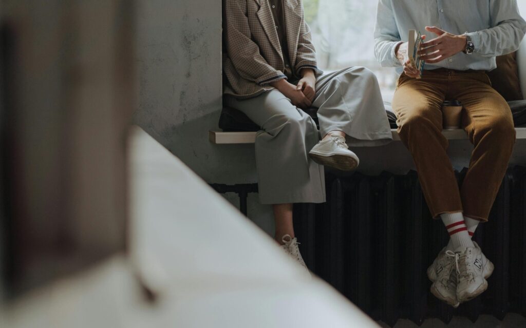 Photo shows two people from the neck down sitting next to each other, slightly facing towards each other. One of them is holing a book.