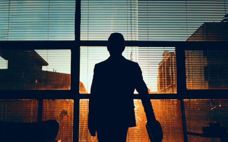 Photo shows the silhouette of a person standing in front of a sunny window, looking towards the light.