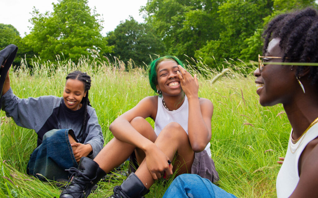 Three young Black people sit together outside in tall grass, smiling, laughing, and talking to one another