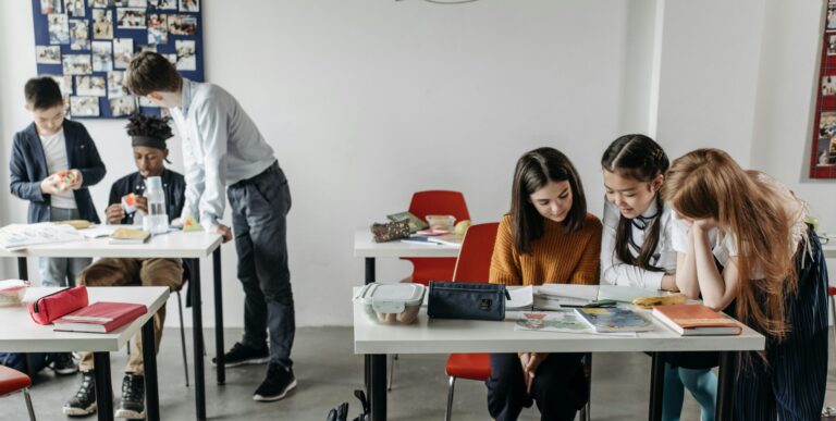A photo of a classroom. An adult stands over a desk speaking to two students. Another group of three students talk to one another and look at a notebook.