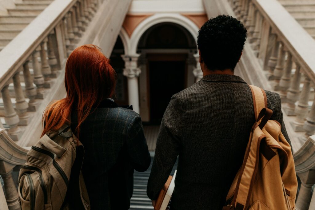 Two students walk in a campus building, facing away from the camera. One has short black hair, and the other has bright red straight hair.