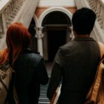 Two students walk in a campus building, facing away from the camera. One has short black hair, and the other has bright red straight hair.