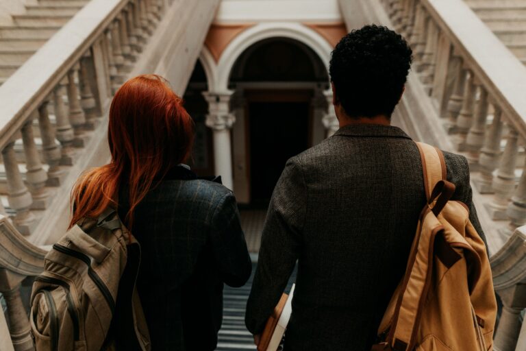 Two students walk in a campus building, facing away from the camera. One has short black hair, and the other has bright red straight hair.