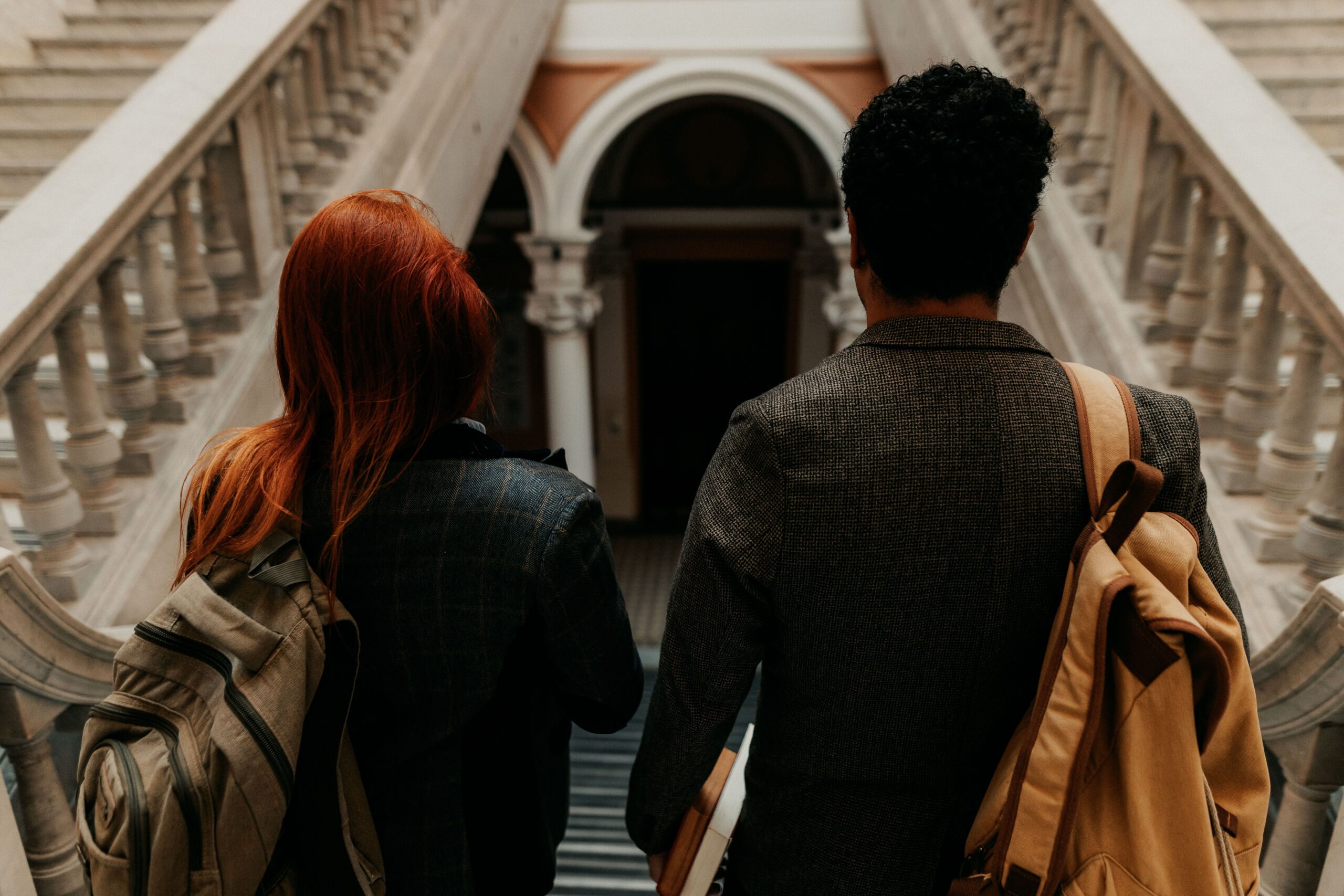 Two students walk in a campus building, facing away from the camera. One has short black hair, and the other has bright red straight hair.