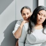Photo of two women with serious expression of their face, standing near a window.