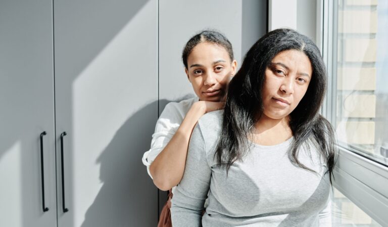Photo of two women with serious expression of their face, standing near a window.